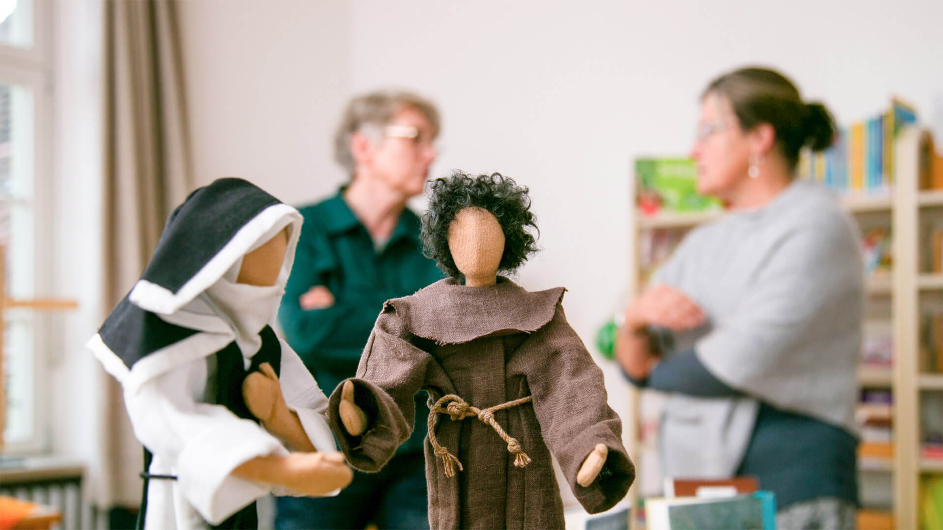 Mönch und Nonne als Puppen im Vordergrund, zwei Frauen unterhalten sich im Hintergrund Mönch und Nonne als Puppen im Vordergrund, zwei Frauen unterhalten sich im Hintergrund